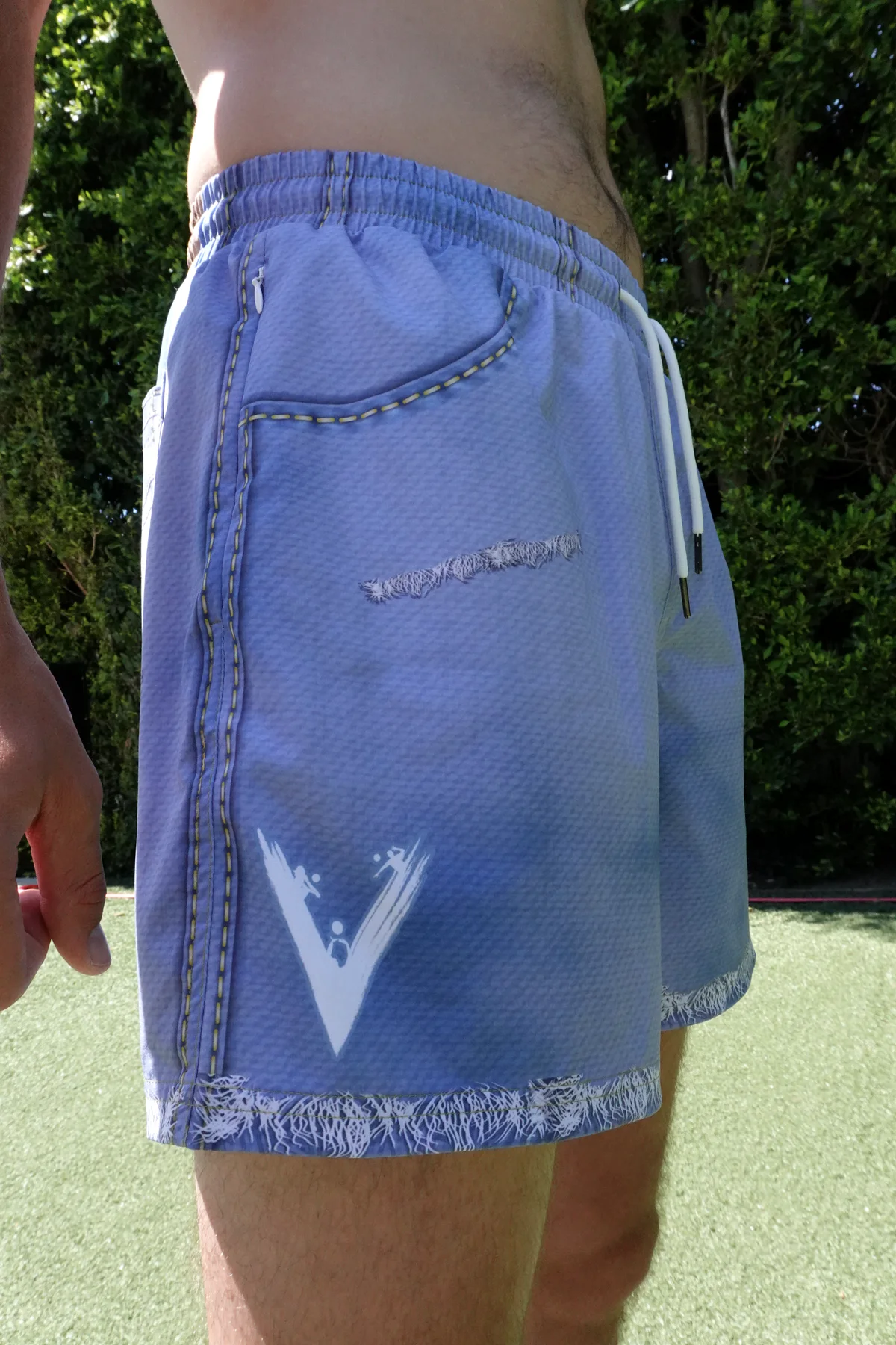 Side fit of JORTS shorts showing the curved pocket, drawcord, V logo, side stitching, and raw hem